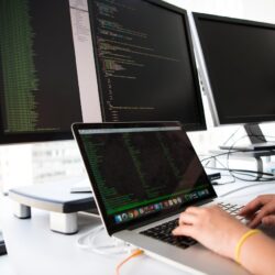 Hands typing on a laptop with 3 additional monitors